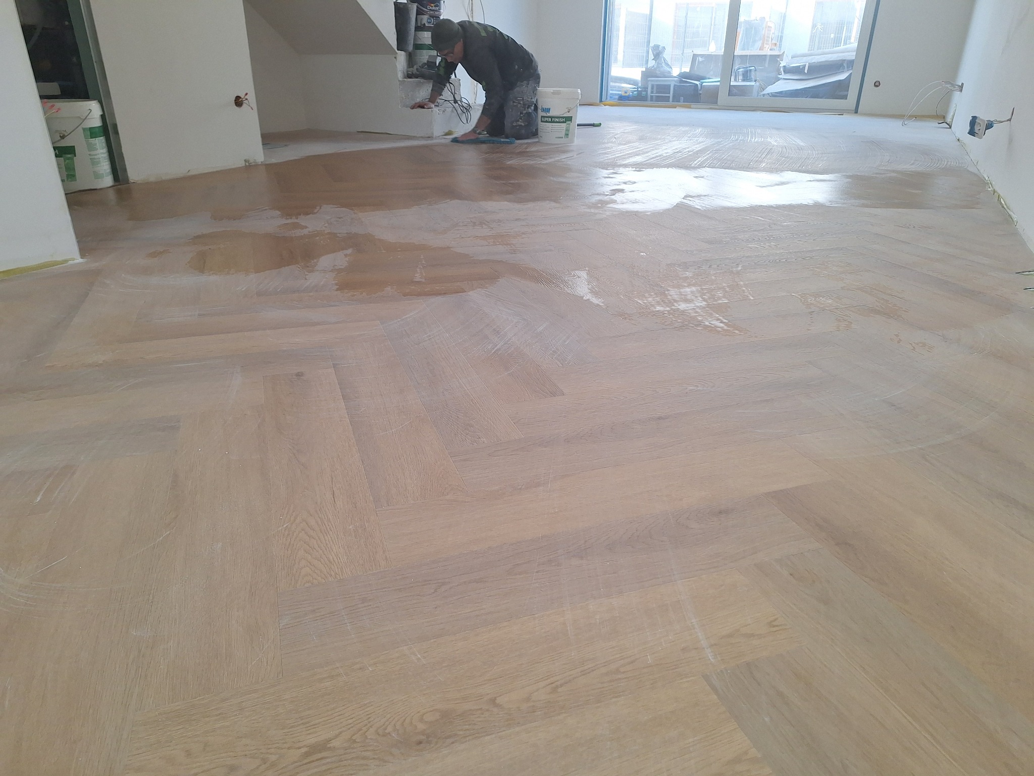 Parkiet w jodełkę zabezpieczany lakierem przez pracownika. Widoczne wiadro z lakierem i narzędzia. Jasne wnętrze z dużym oknem. Etap prac wykończeniowych.