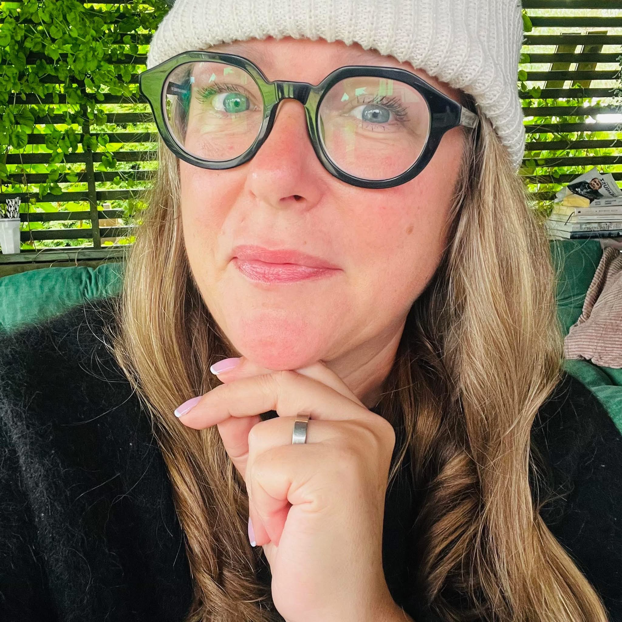 Portret kobiety w okularach i czapce, z dłońmi pod brodą, na tle zielonej roślinności i książek. Ujęcie z bliska, z lekkim uśmiechem i spojrzeniem w obiektyw.