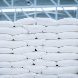 EMPRO MARCIN RYTEL - Środki Do Produkcji Rolnej - Stos białych worków ułożonych warstwami w magazynie. Widoczne szwy i zawiązania worków. Metalowa konstrukcja dachu w tle. Delikatne oświetlenie.