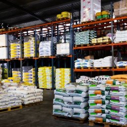 EMPRO MARCIN RYTEL - Środki Do Produkcji Rolnej - Magazyn z paletami worków na regałach. Różne kolory i rodzaje opakowań. Widok na pełne półki z zapasami. Oświetlenie magazynowe.