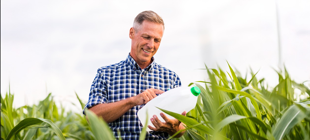 Uśmiechnięty rolnik w polu kukurydzy, trzymający biały kanister z zieloną nakrętką, prawdopodobnie z płynnym nawozem. Kontrola jakości lub przygotowanie do użycia.