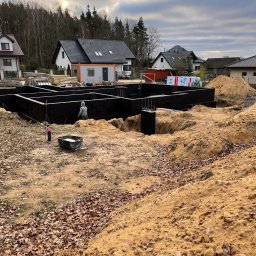 HOLBud - Fundamenty domu jednorodzinnego w trakcie izolacji bitumicznej, widoczne zbrojenia i wykop. Prace ziemne w toku, w tle zabudowa jednorodzinna i las.