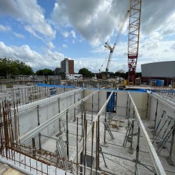 HOLBud - Wysoki kadr z budowy fundamentów: szalunki, zbrojenie, podpory i żuraw na tle błękitnego nieba z chmurami. Widoczne budynki w oddali.