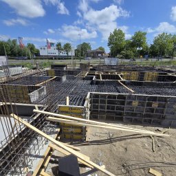 HOLBud - Szeroki kadr na zbrojenie fundamentów z żółtymi elementami szalunkowymi, widoczne niebo i budynki w tle. Ujęcie z góry, podkreślające rozległość prac.