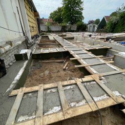 HOLBud - Wykop pod fundamenty z częściowo wylanym betonem, szalunki z desek, widoczne zbrojenia. Prace budowlane w toku, widok z góry na plac budowy i fragment budynku w tle.