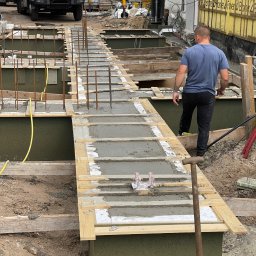 HOLBud - Wykop fundamentowy z częściowo wylanym betonem, widoczne zbrojenia i szalunki. W tle betoniarka i pracownik budowlany. Ujęcie z perspektywy ziemi.