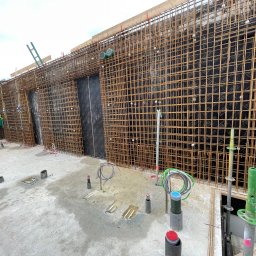 HOLBud - Siatka zbrojeniowa na czarnej membranie, widok z poziomu wylewki. Elementy instalacji elektrycznej wystają z betonu. Szalunek z desek w tle.