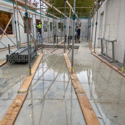 HOLBud - Wnętrze budynku żelbetowego w trakcie budowy, widoczne szalunki, podpory i pracownicy na rusztowaniach. Podłoże mokre, odbijające elementy konstrukcji.