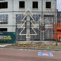 HOLBud - Surowy budynek modułowy w trakcie budowy, ozdobiony świątecznymi, neonowymi dekoracjami w kształcie choinki i renifera. Widoczny podnośnik koszowy i baner informacyjny.