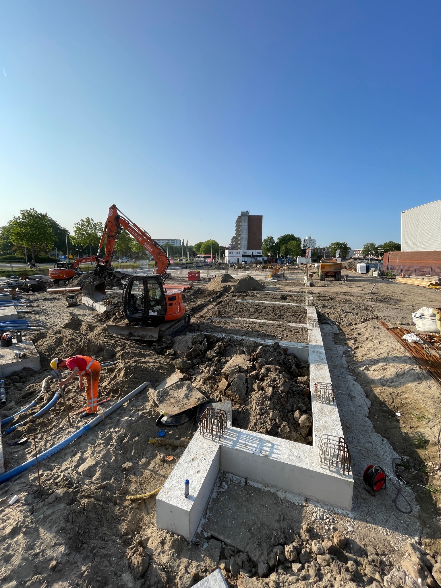 Budowa fundamentów: wykop, częściowo zalane betonem zbrojenia, pracownik w kasku i koparka Hitachi na placu budowy w słoneczny dzień.