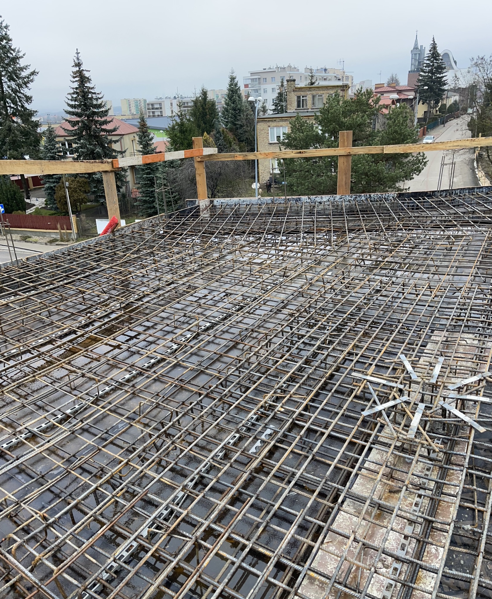Siatka zbrojeniowa na mokrej powierzchni, drewniane podpory, w tle zabudowa miejska i drzewa. Widok z góry na konstrukcję z metalowych prętów.