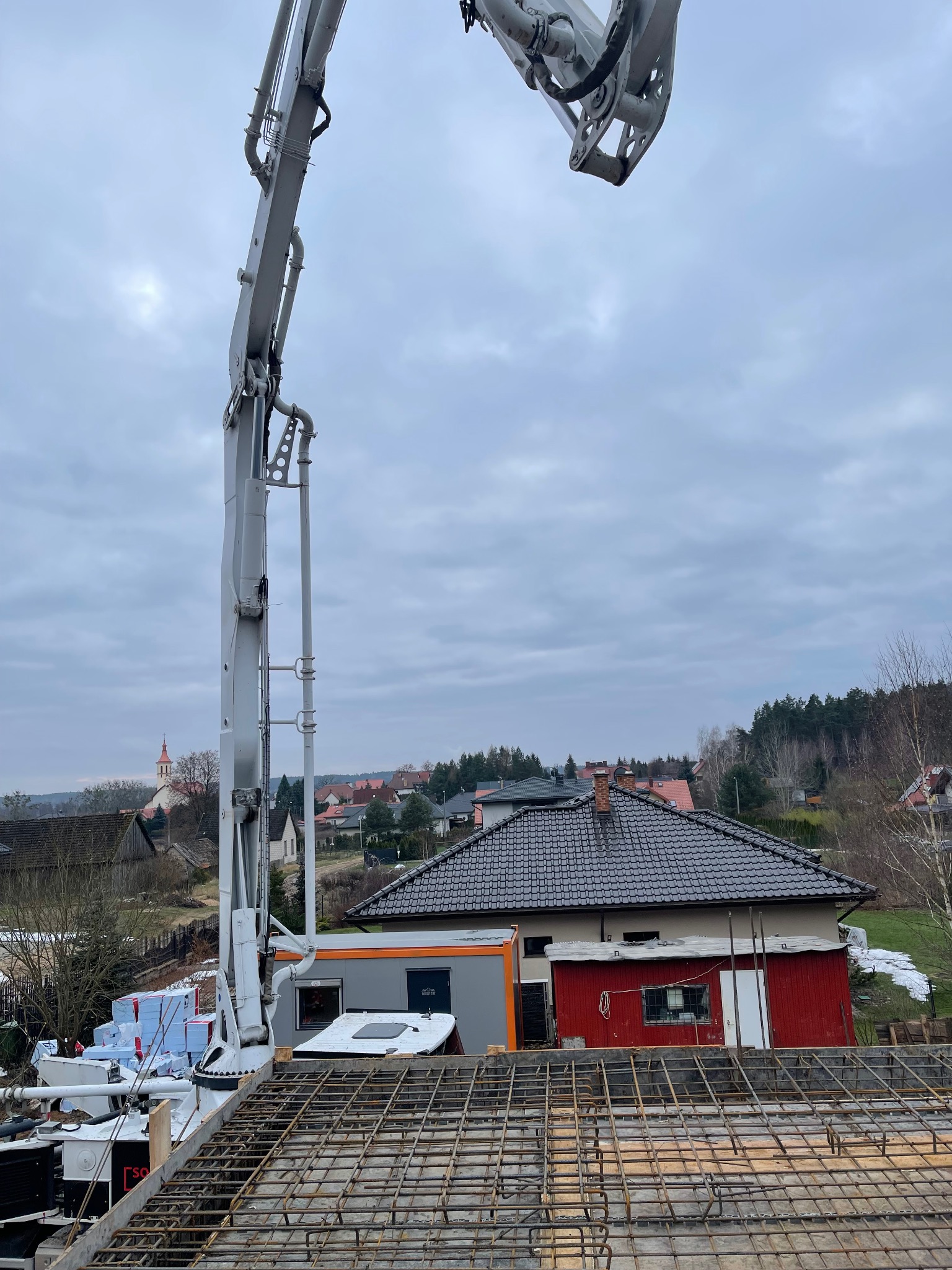 Zbrojenie stropu domu jednorodzinnego z widoczną pompą do betonu na tle zabudowy wiejskiej i pochmurnego nieba. Widok z góry na siatkę zbrojeniową.
