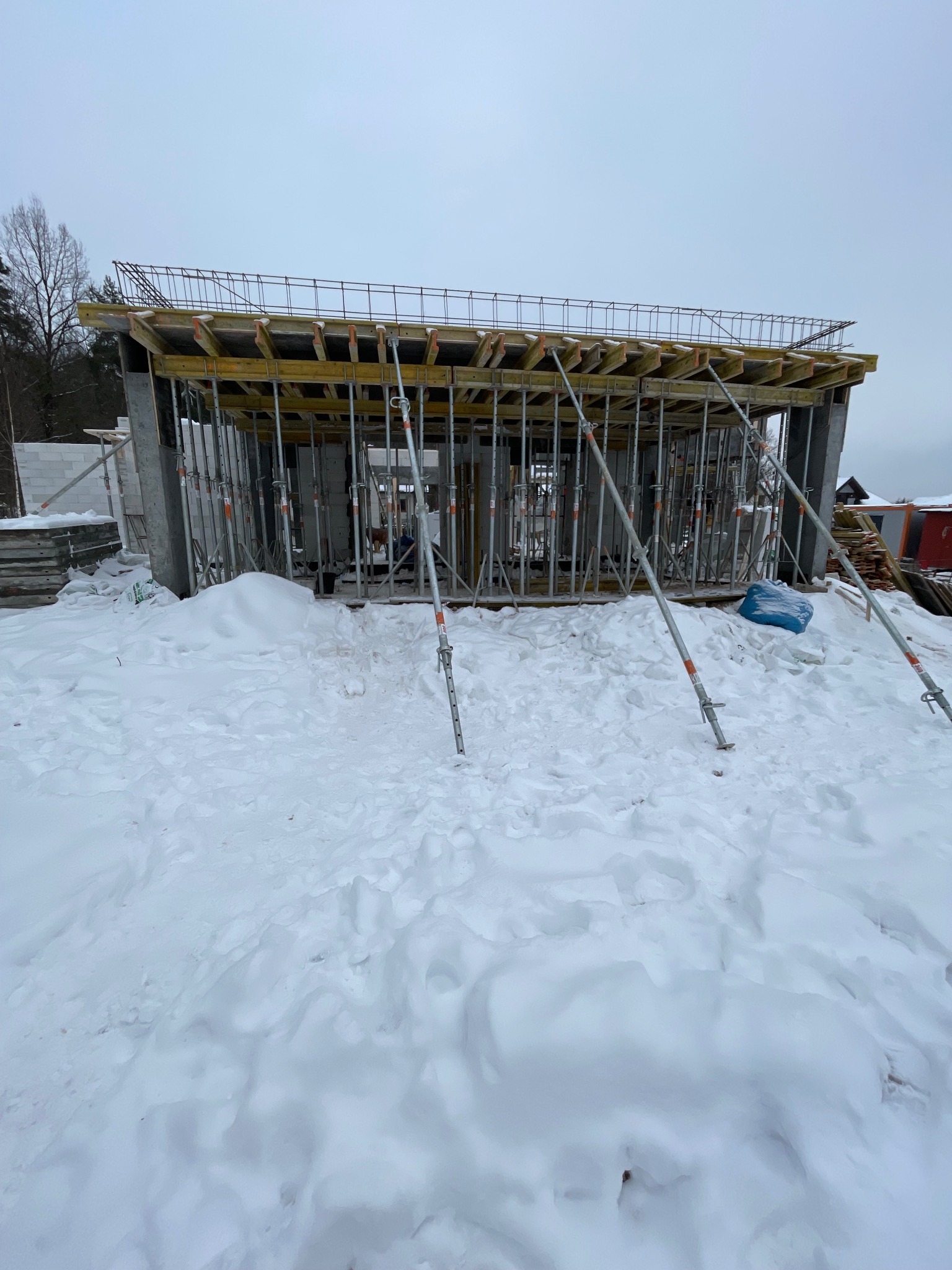 Surowy stan budynku żelbetowego w zimowej scenerii, widoczne szalunki i podpory, teren pokryty śniegiem, pochmurne niebo w tle.