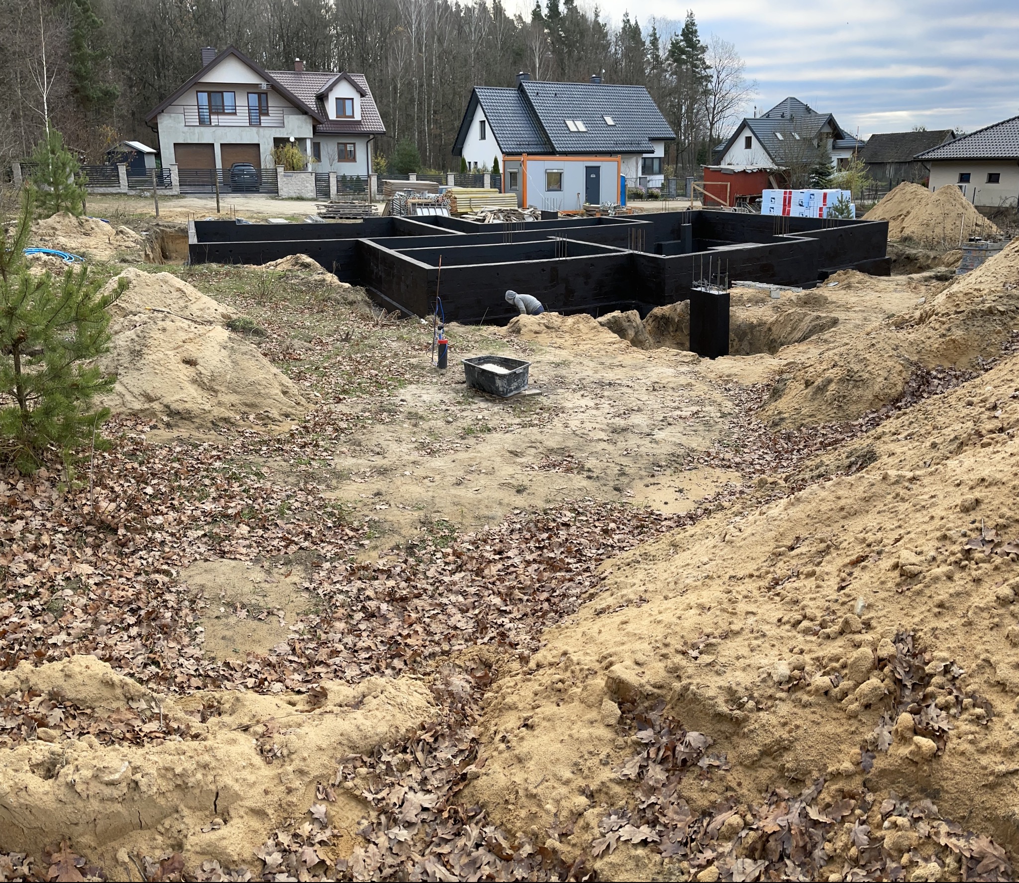 Czarne, zaizolowane fundamenty domu jednorodzinnego w wykopie, z widocznymi prętami zbrojeniowymi. W tle zabudowa mieszkalna i drzewa.