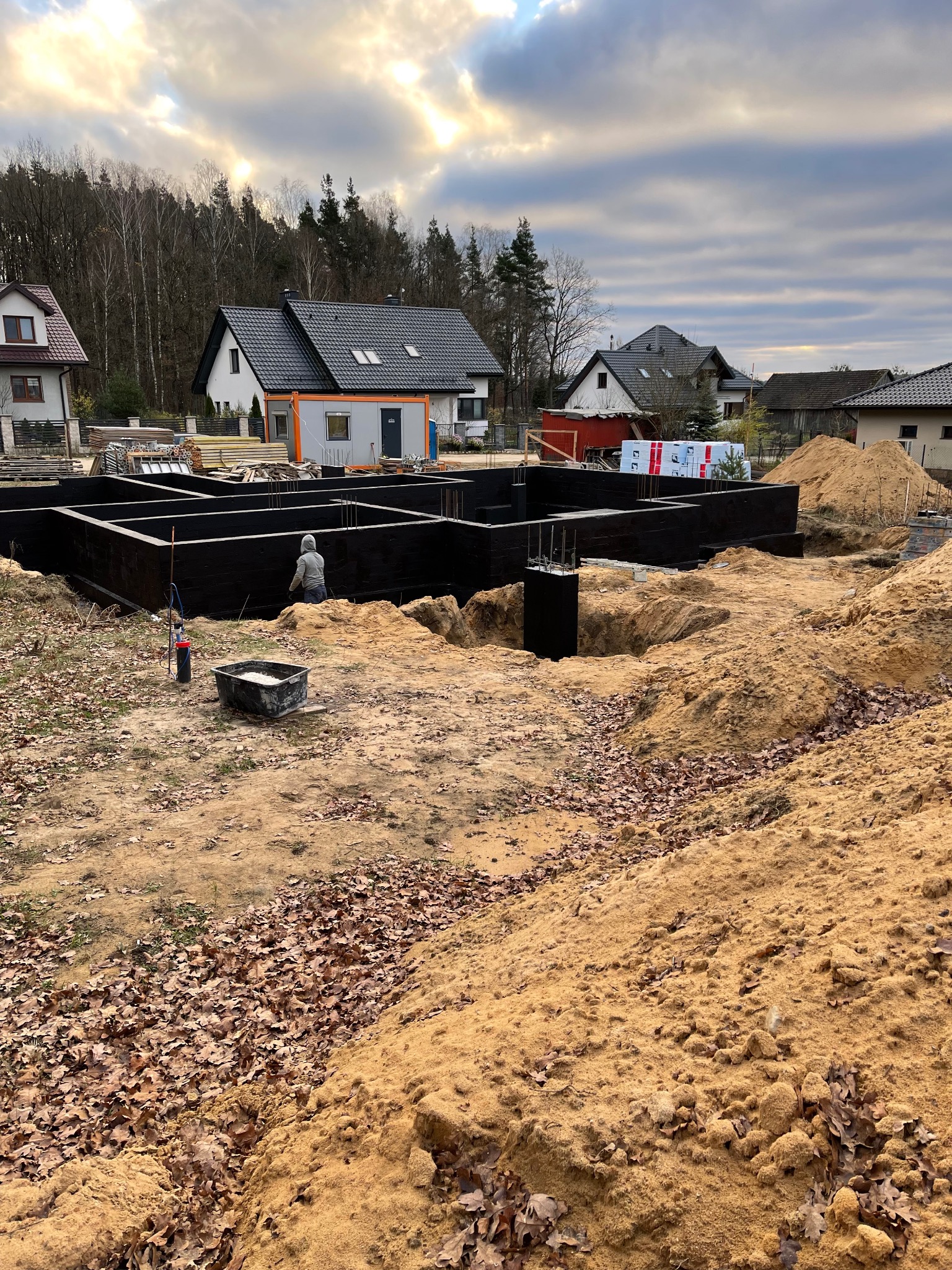 Fundamenty domu jednorodzinnego w trakcie izolacji bitumicznej, widoczne zbrojenia i wykop. Prace ziemne w toku, w tle zabudowa jednorodzinna i las.