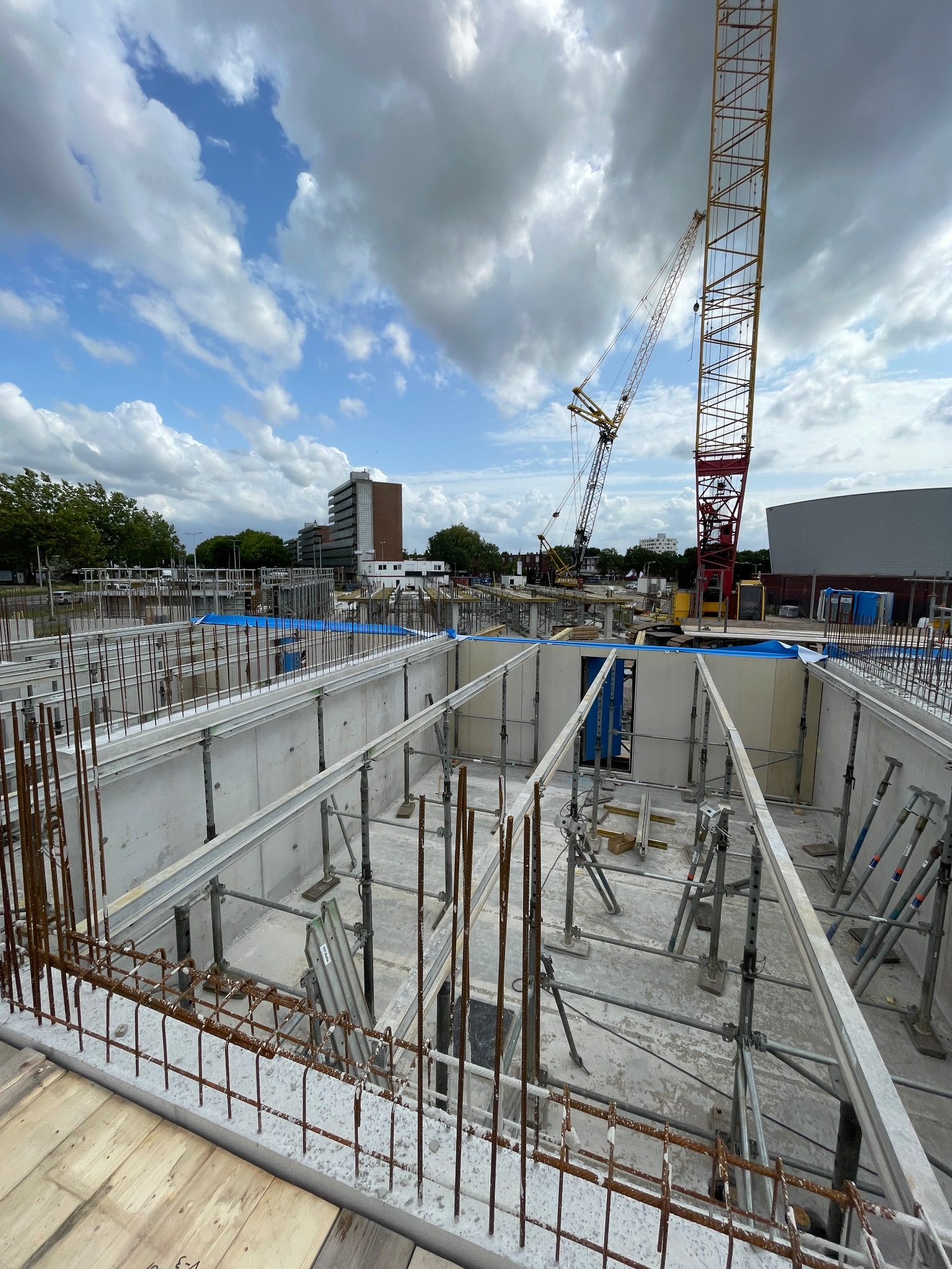 Wysoki kadr z budowy fundamentów: szalunki, zbrojenie, podpory i żuraw na tle błękitnego nieba z chmurami. Widoczne budynki w oddali.