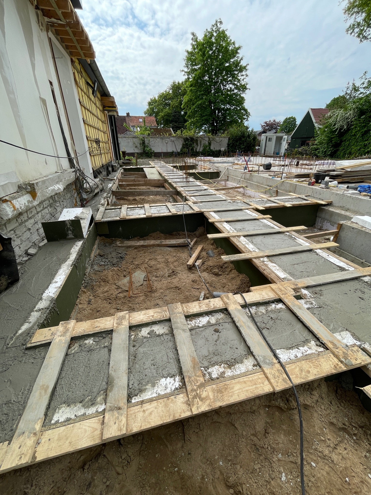 Wykop pod fundamenty z częściowo wylanym betonem, szalunki z desek, widoczne zbrojenia. Prace budowlane w toku, widok z góry na plac budowy i fragment budynku w tle.