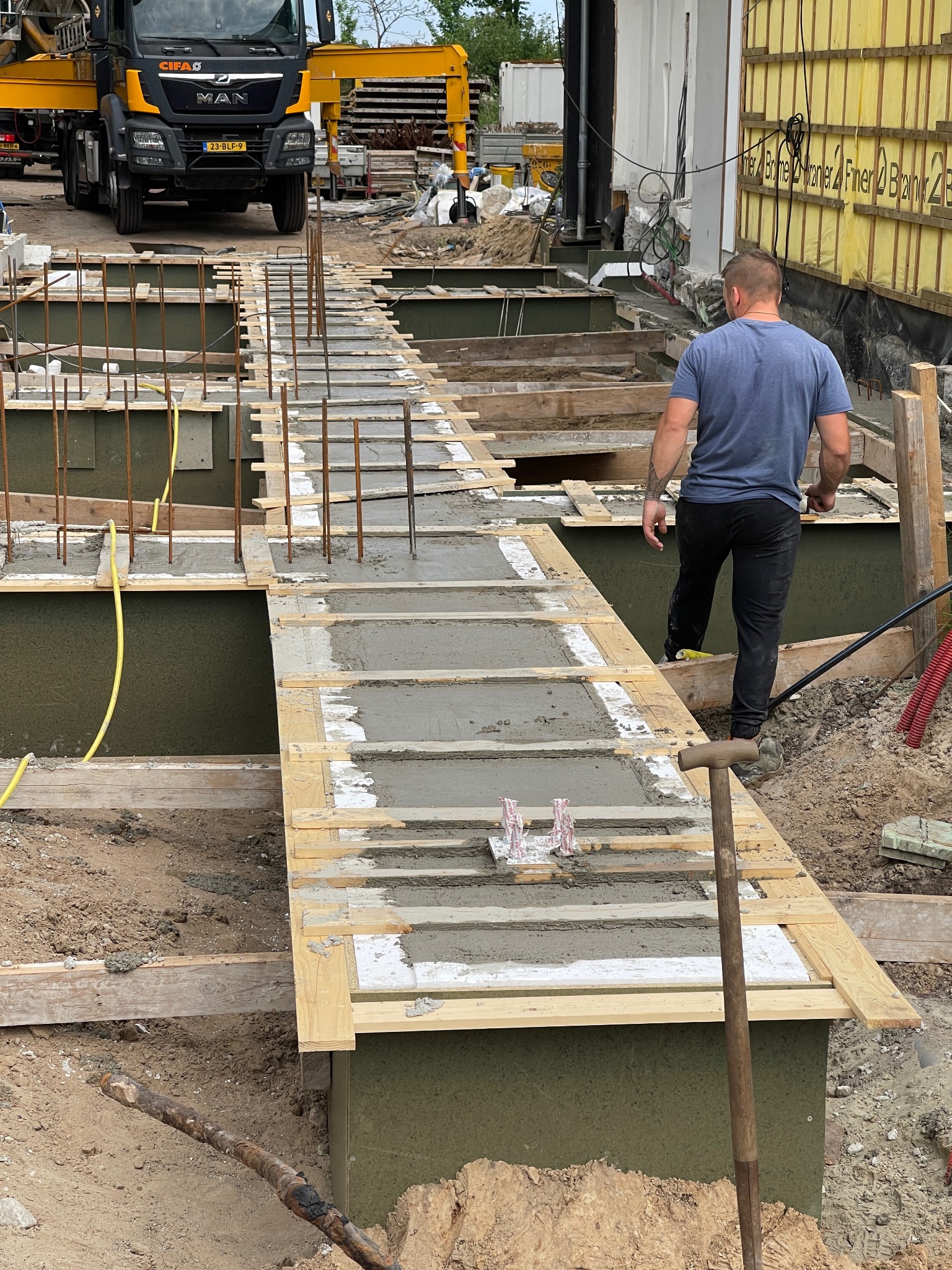 Wykop fundamentowy z częściowo wylanym betonem, widoczne zbrojenia i szalunki. W tle betoniarka i pracownik budowlany. Ujęcie z perspektywy ziemi.