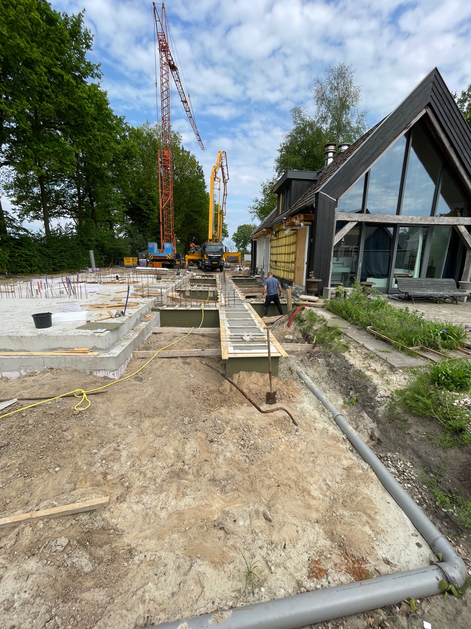 Budowa domu: fundamenty zbrojone, betonowe, częściowo zalane. Na drugim planie dźwig i pompa do betonu. W tle nowoczesny dom z dużymi oknami.