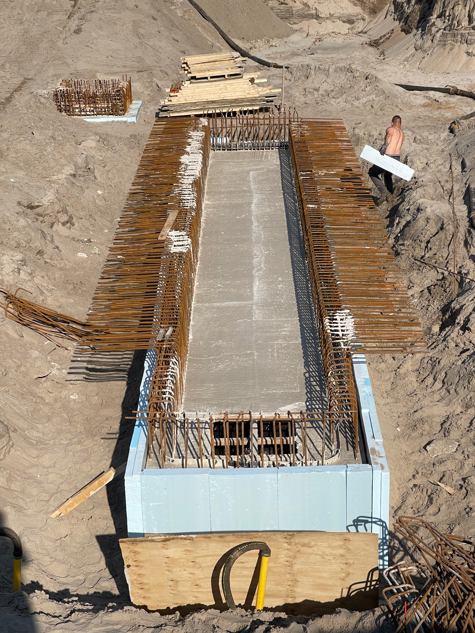 Z góry: Zalany beton w szalunku z izolacją i zbrojeniem. Na piasku leżą elementy zbrojenia i drewniane deski. W tle pracownik budowlany przenosi izolację.