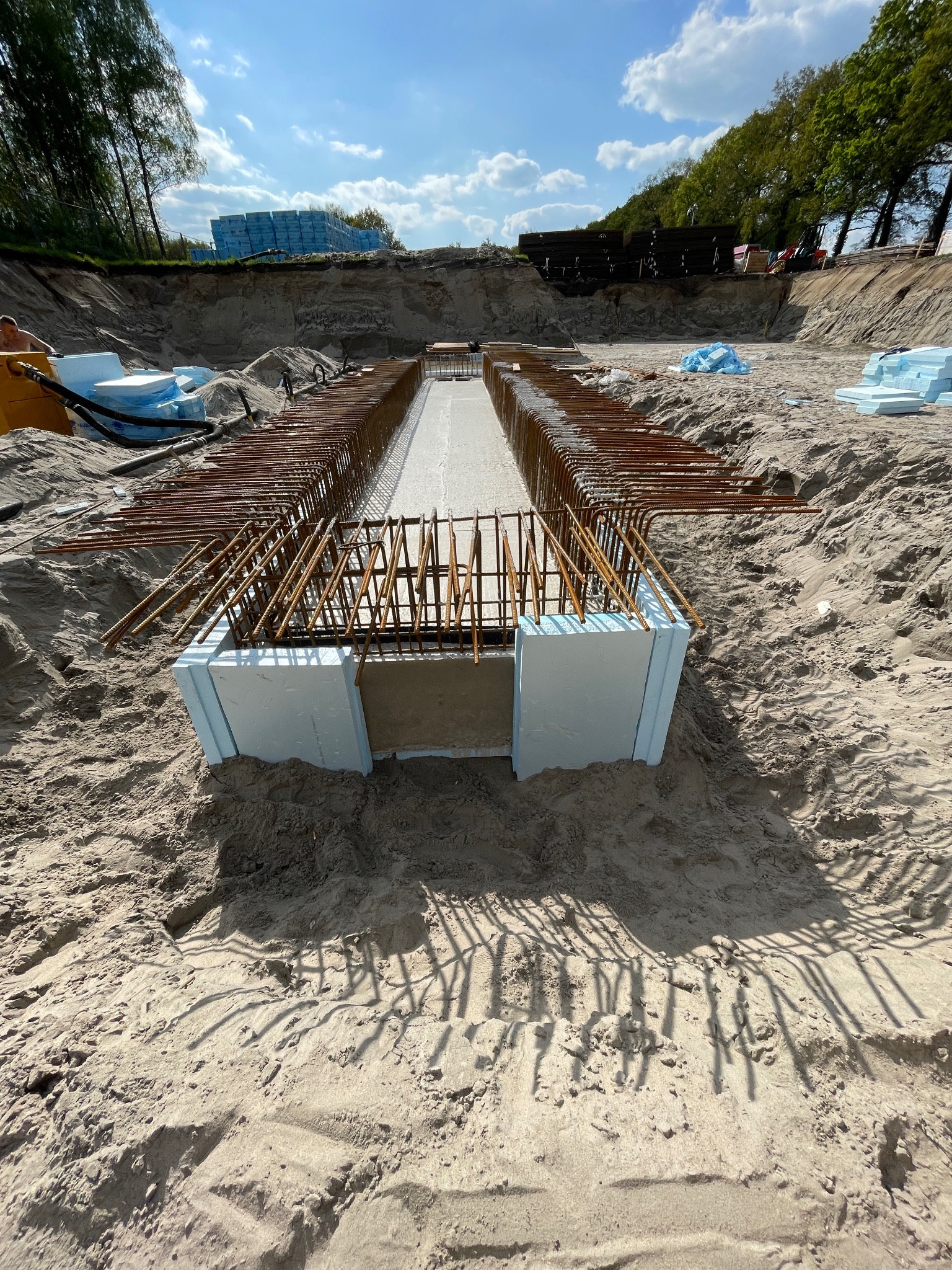 Zbrojenie fundamentu z izolacją termiczną w wykopie. Widoczne stalowe pręty i białe płyty izolacyjne. W tle ziemia, drzewa i błękitne niebo.