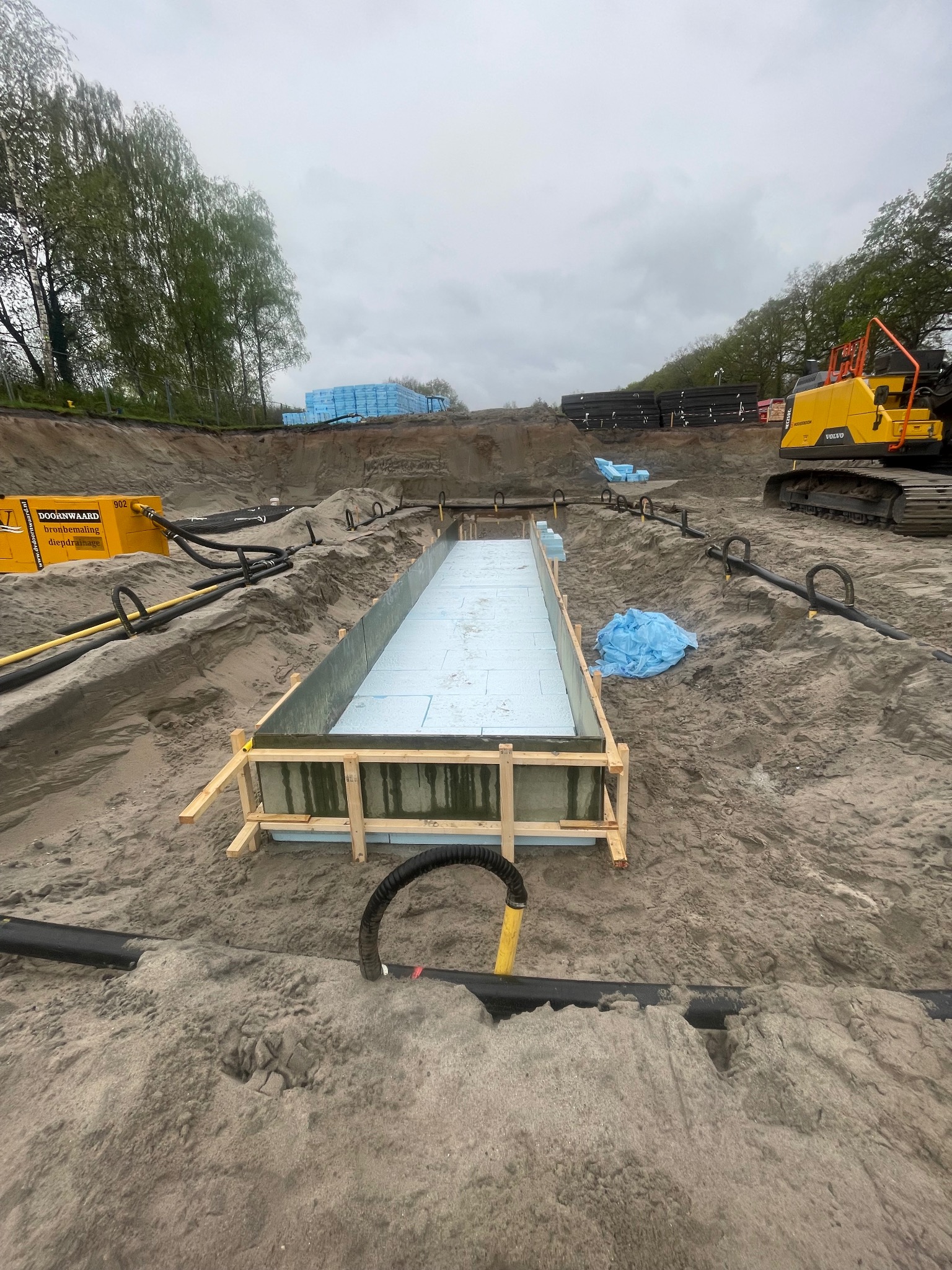 Wykop pod fundamenty z drewnianym szalunkiem i izolacją z pianki. W tle widoczna koparka Volvo i materiały budowlane. Prace ziemne w toku.