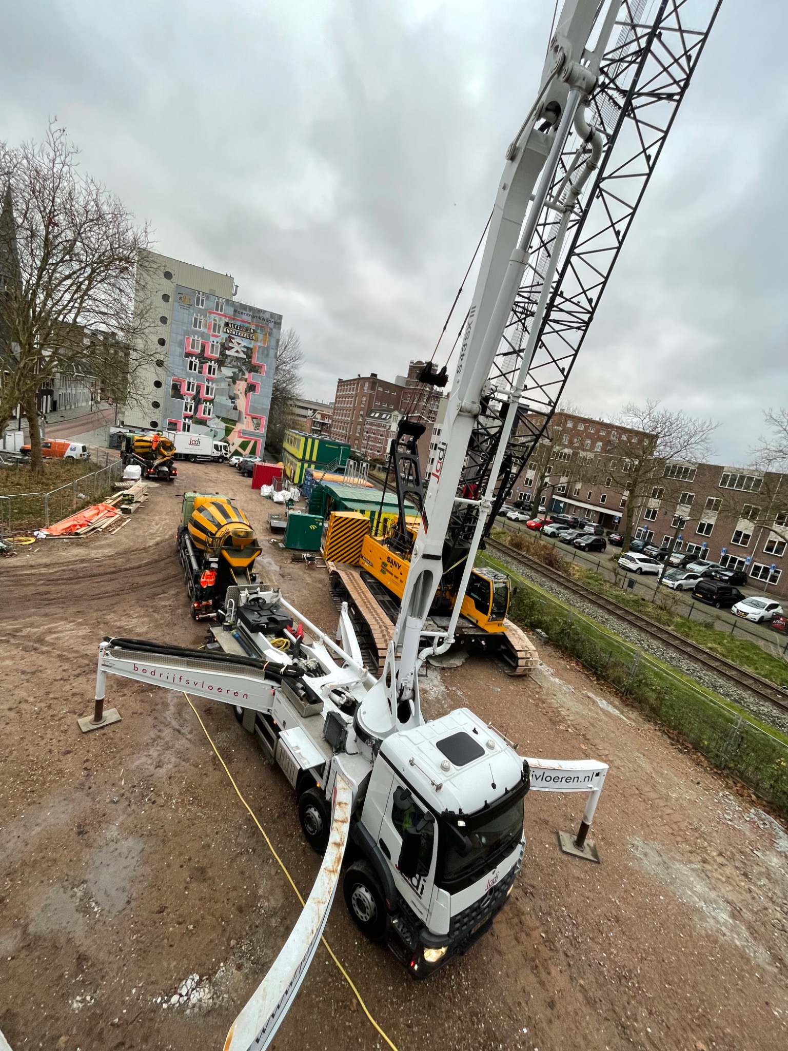 Betoniarka i dźwig na placu budowy w pochmurny dzień. Widok z góry ukazuje maszyny, budynki w tle i fragment torów kolejowych. Na dźwigu widoczne logo firmy.