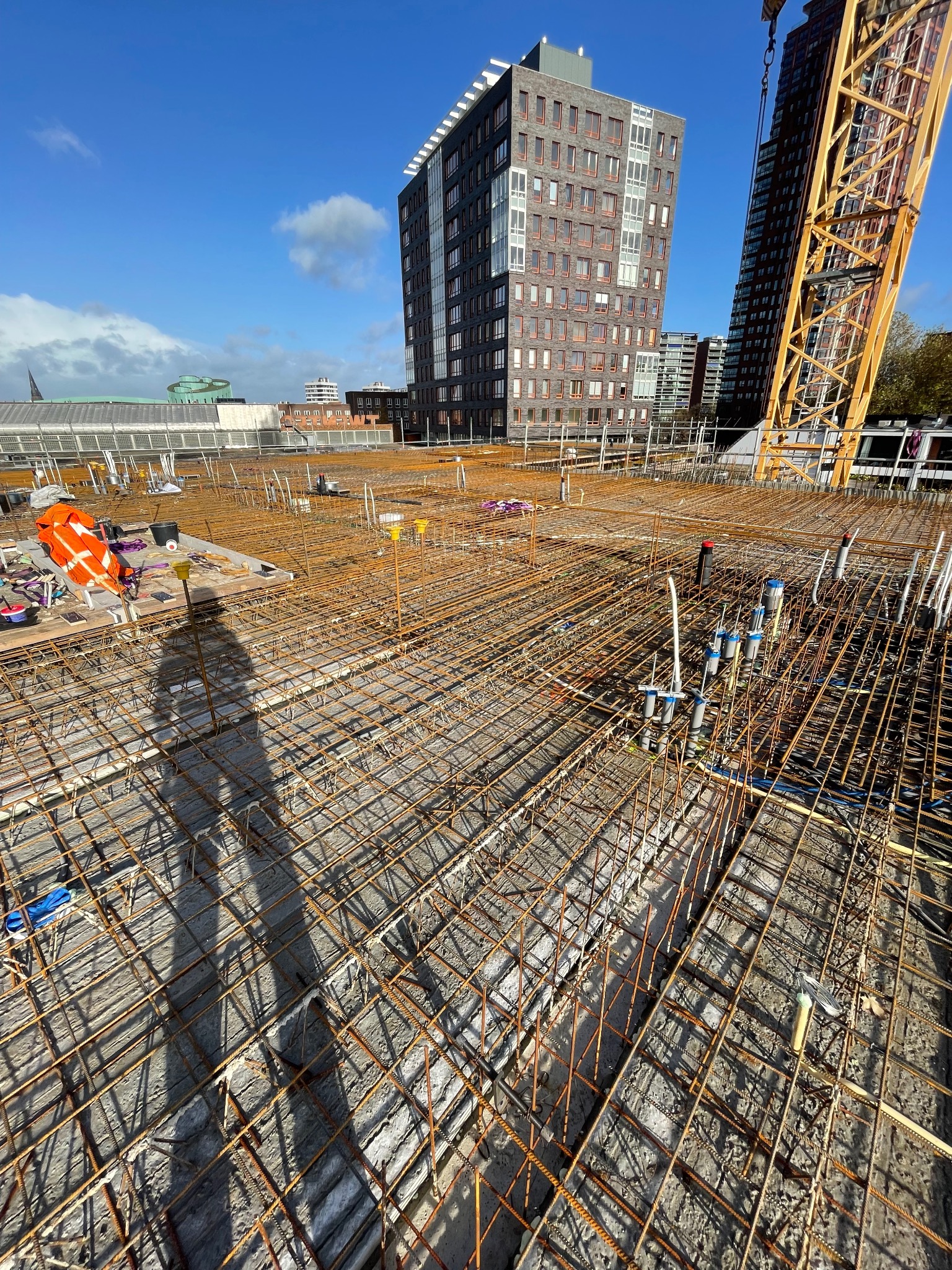 Siatka zbrojeniowa na budowie, widok z góry, z shadow sylwetki osoby. W tle budynki miejskie i żuraw budowlany. Elementy instalacji pionowej wystają z betonu.