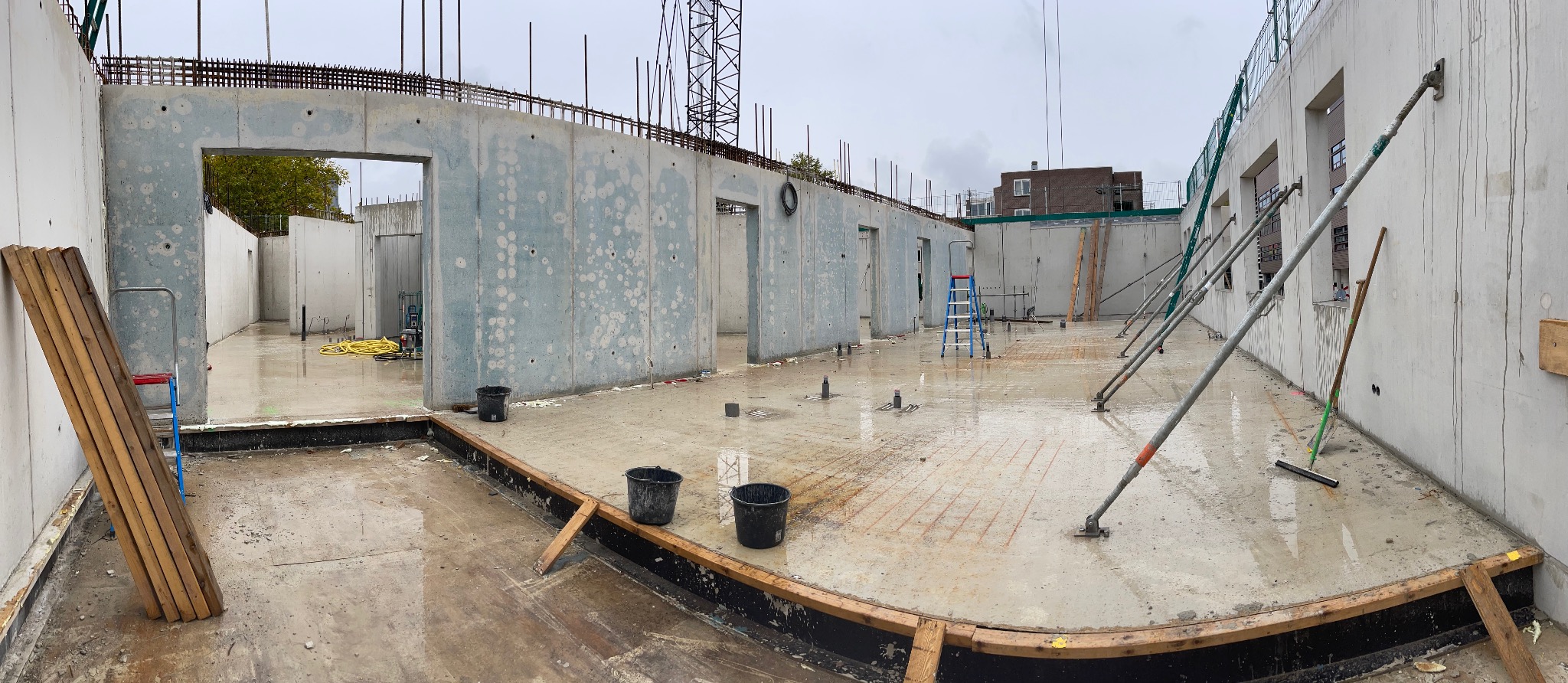 Wnętrze budynku żelbetowego w trakcie budowy. Widoczne ściany z otworami okiennymi i drzwiowymi, zbrojenie na górze ścian, podpory i narzędzia budowlane.