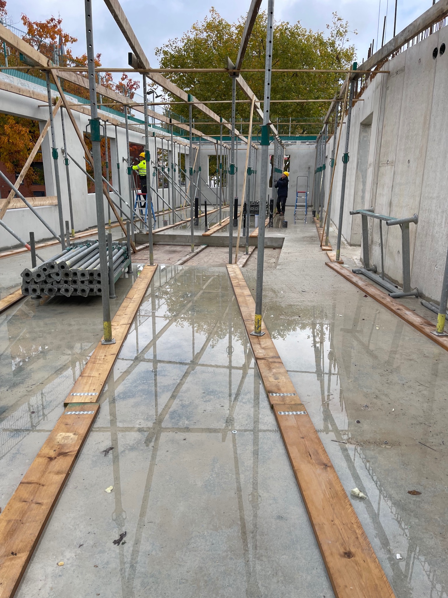 Wnętrze budynku żelbetowego w trakcie budowy, widoczne szalunki, podpory i pracownicy na rusztowaniach. Podłoże mokre, odbijające elementy konstrukcji.