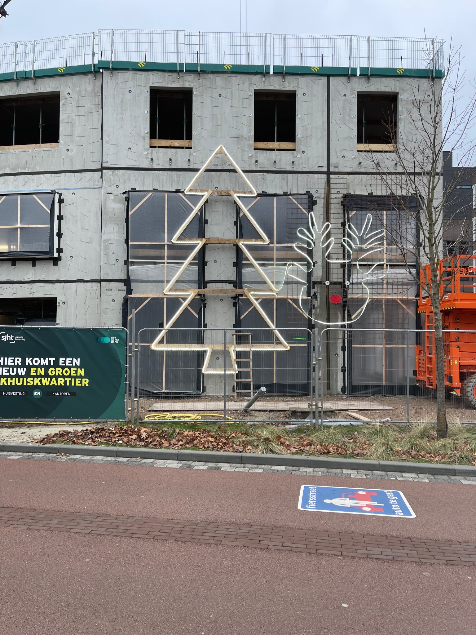 Surowy budynek modułowy w trakcie budowy, ozdobiony świątecznymi, neonowymi dekoracjami w kształcie choinki i renifera. Widoczny podnośnik koszowy i baner informacyjny.