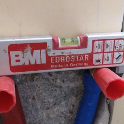 robcioinstalacjesanitarne - Poziomica BMI Eurostar na tle ściany z wystającymi rurami (czerwona i niebieska) w bruździe. Widoczne oznaczenia na poziomicy.