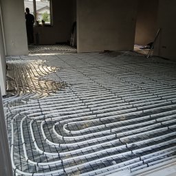 robcioinstalacjesanitarne - Białe rury ogrzewania podłogowego ułożone w regularnych pętlach na folii izolacyjnej w jasnym pomieszczeniu. Widoczny fragment okna i sylwetka osoby w tle.