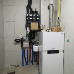 Instalacje sanitarne - Nowoczesny piec Viessmann z instalacją grzewczą w kotłowni wykończonej szarą płytką. Widoczne rury, zawory, filtry i elementy sterujące.