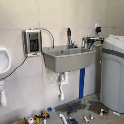 Hydraulik - Instalacje Sanitarne - Instalacja hydrauliczna: umywalka przemysłowa, pisuar i zmiękczacz wody na tle kafelkowanej ściany. Widoczne narzędzia i elementy instalacyjne na podłodze.