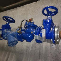 Hydraulik - Instalacje Sanitarne - Niebieskie zawory przemysłowe z manometrem, zamontowane na rurach przy szarej, surowej ścianie. Widoczne elementy mocujące i fragment instalacji pod sufitem.
