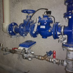 Hydraulik - Instalacje Sanitarne - Niebieska instalacja wodna z zaworami i czujnikiem przepływu na szarej ścianie. Różne rury i złącza tworzą skomplikowany system. Widoczne ślady użytkowania.
