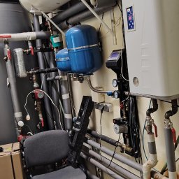 Hydraulik - Instalacje Sanitarne - Rozbudowana instalacja grzewcza i wodna z poziomym bojlerem, dwoma niebieskimi naczyniami przeponowymi, białym piecem i licznymi rurami na żółtej ścianie, obok szare krzesło i poziomica.