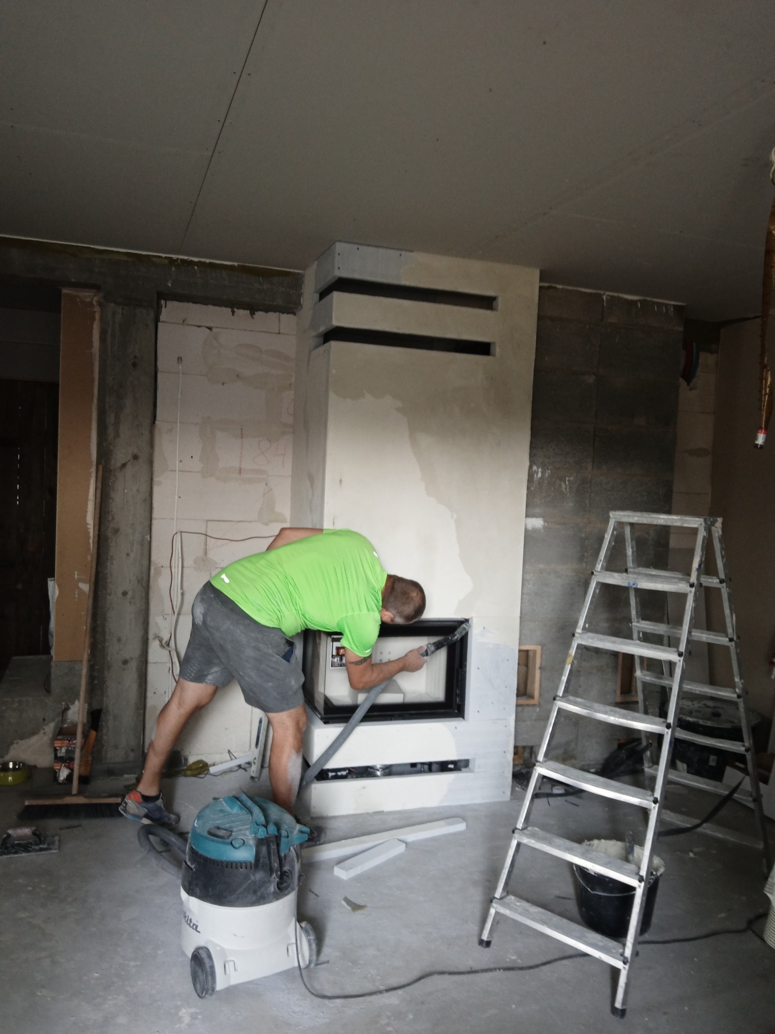 Mężczyzna w zielonej koszulce wygładza szpachelką obudowę kominka w surowym wnętrzu. Widoczny odkurzacz przemysłowy i drabina. Prace wykończeniowe w toku.