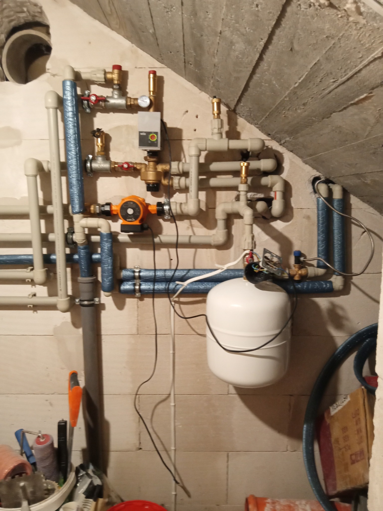 Złożona instalacja grzewcza z rur, zaworów i pompy obiegowej w pomieszczeniu z surowym betonowym stropem. Widoczne narzędzia i akcesoria instalacyjne.