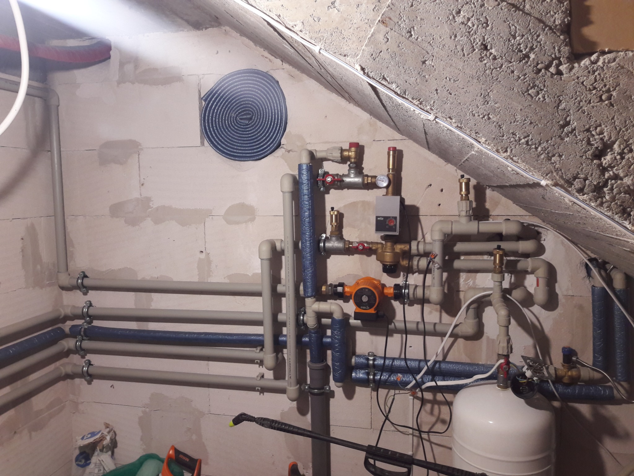 Złożona instalacja grzewcza z rurami, zaworami i pompą obiegową na tle surowej ściany z betonu komórkowego i zwiniętej izolacji. Widoczne elementy montażowe i przyłączeniowe.