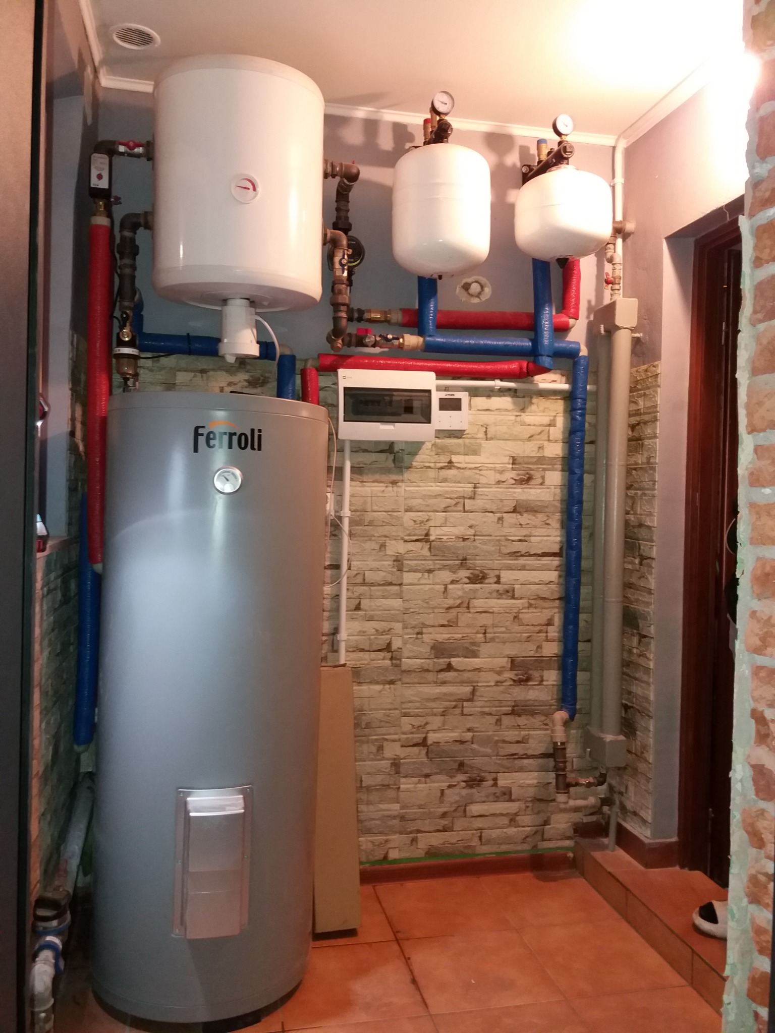 Szary piec Ferroli z podłączonymi rurami w kolorach czerwonym i niebieskim, na tle ściany z cegły w Białymstoku. Widoczne także białe zbiorniki i instalacja elektryczna.