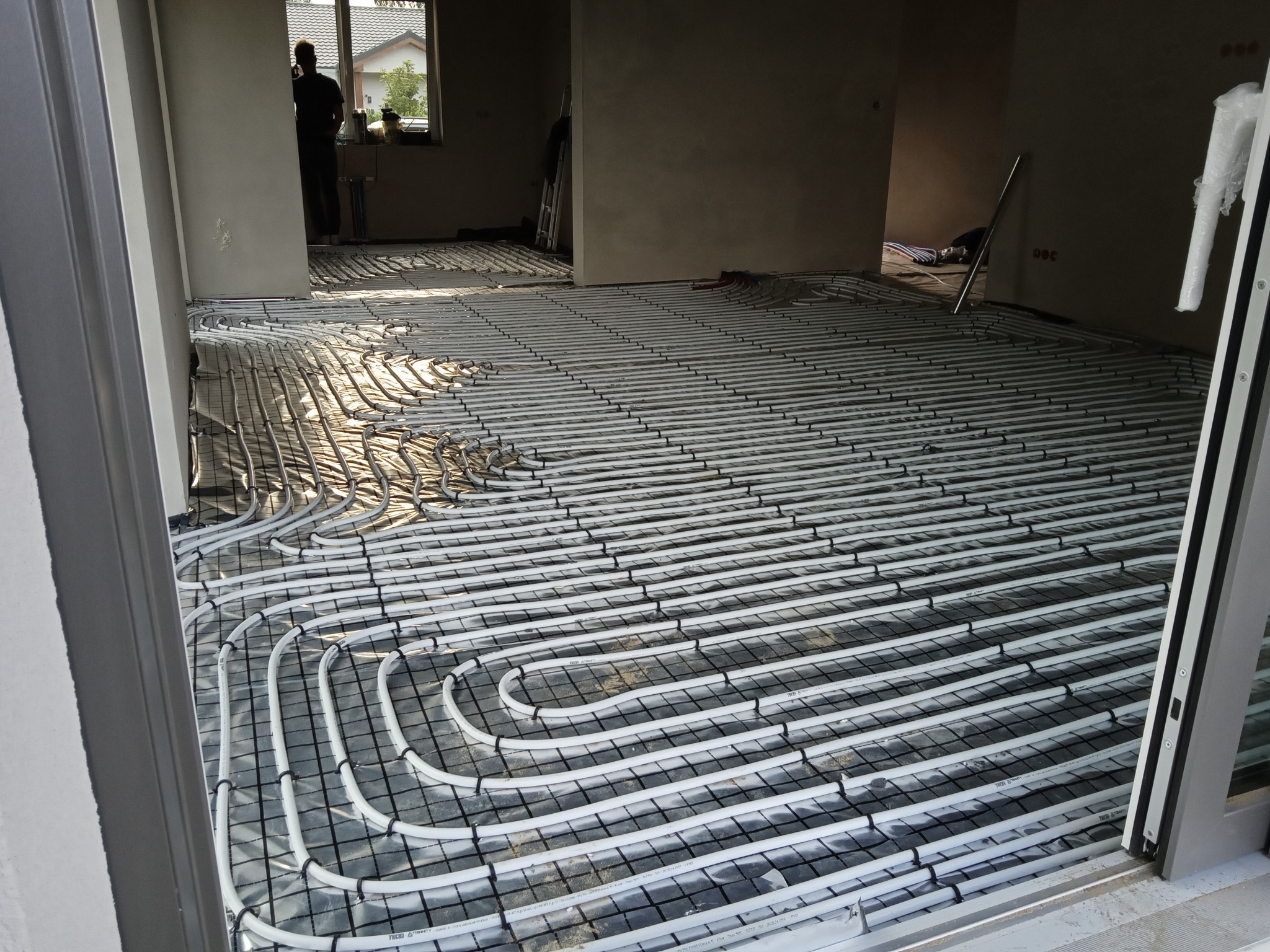 Białe rury ogrzewania podłogowego ułożone w regularnych pętlach na folii izolacyjnej w jasnym pomieszczeniu. Widoczny fragment okna i sylwetka osoby w tle.