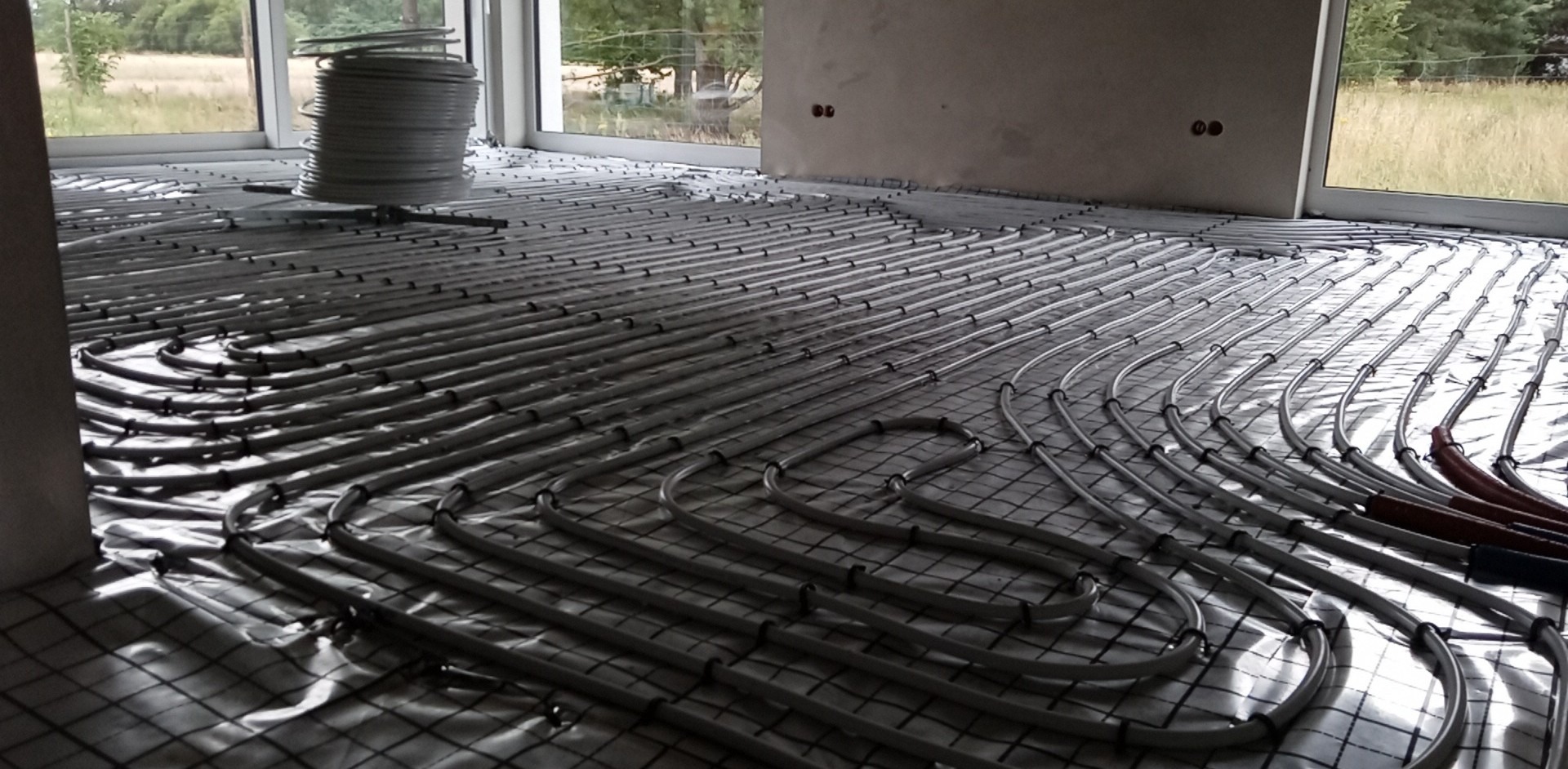 Szare rury ogrzewania podłogowego ułożone na folii izolacyjnej w jasnym pomieszczeniu z dużymi oknami. Widoczny fragment ściany z puszkami elektrycznymi.