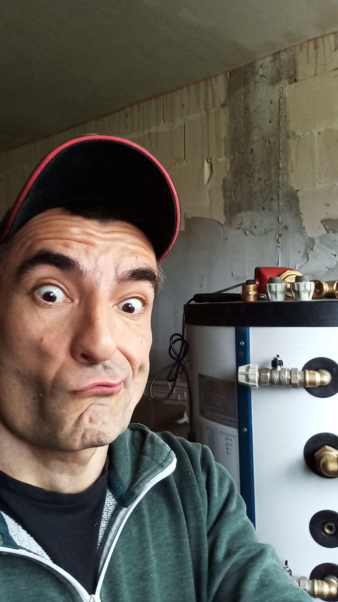 Autoportret hydraulika z bojlerem w surowym wnętrzu. Mężczyzna w czapce z czerwonym daszkiem robi zabawną minę, a za nim widoczny biały bojler z licznymi przyłączami.