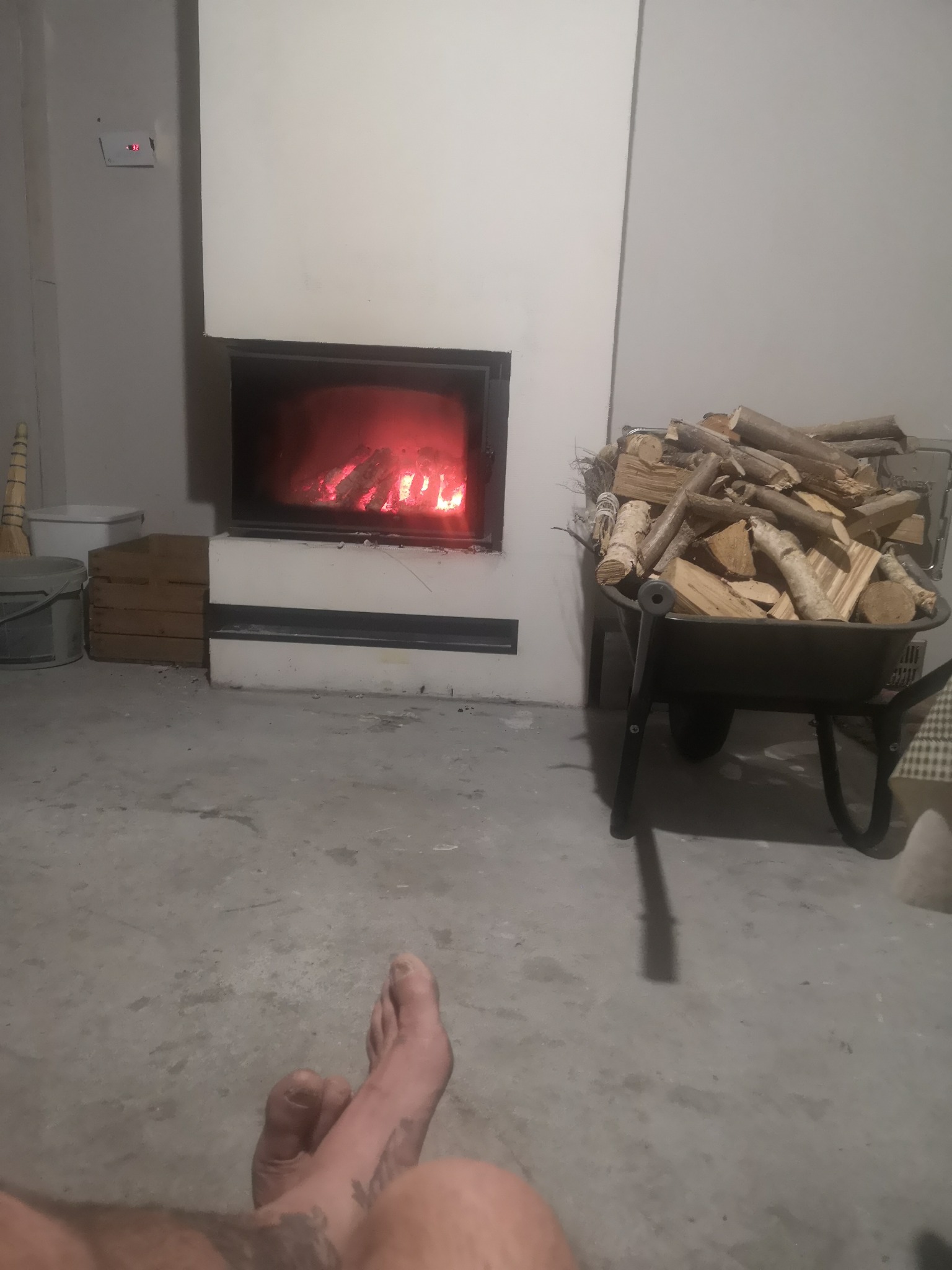 Nowoczesny kominek z paleniskiem, obok taczka z drewnem. Widoczne stopy osoby siedzącej na podłodze. Minimalistyczny design, surowe wykończenie wnętrza.