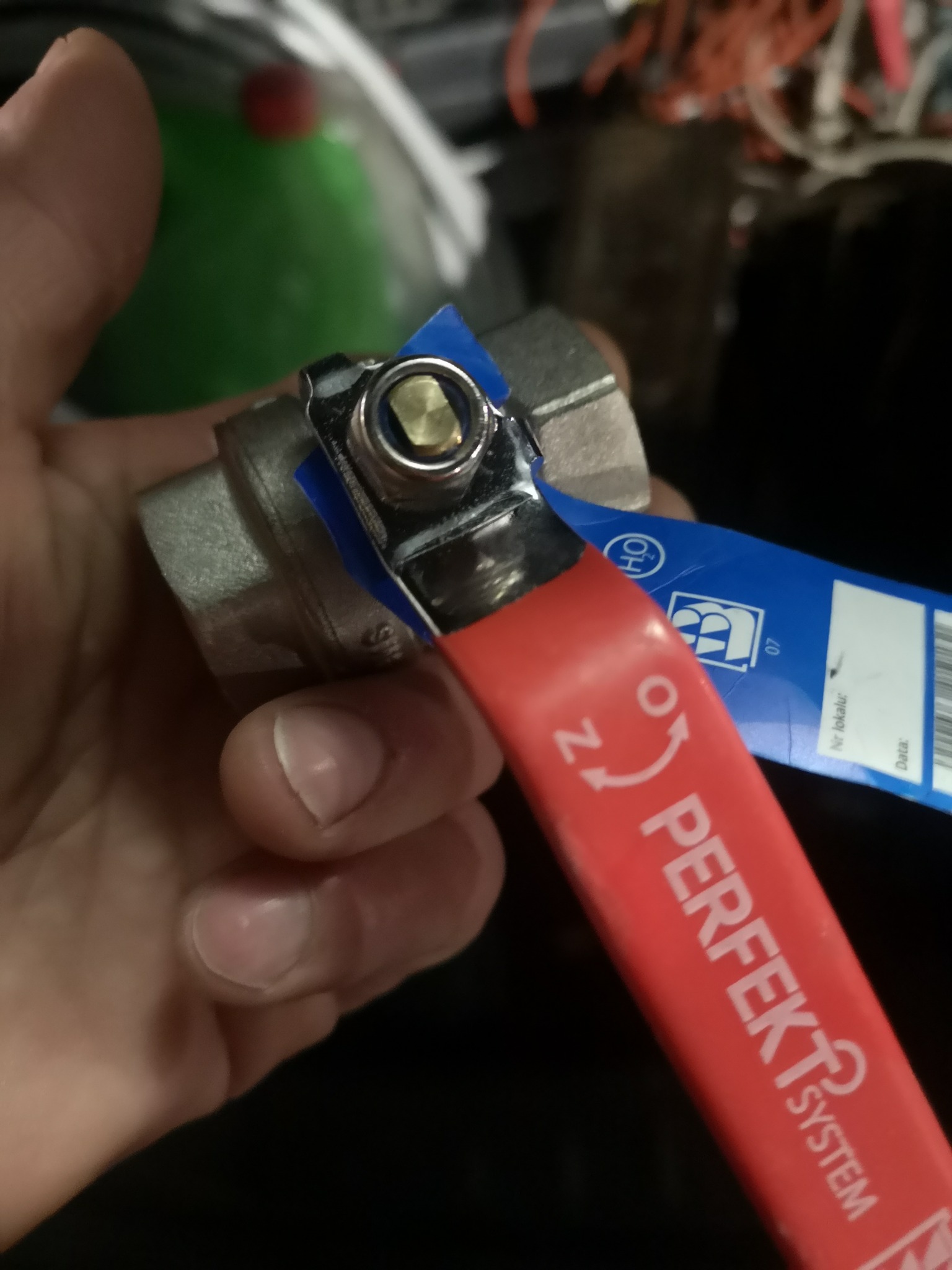 Zawór kulowy z czerwoną rączką marki Perfekt System, trzymany w dłoni, z widoczną niebieską etykietą. Element instalacji hydraulicznej.