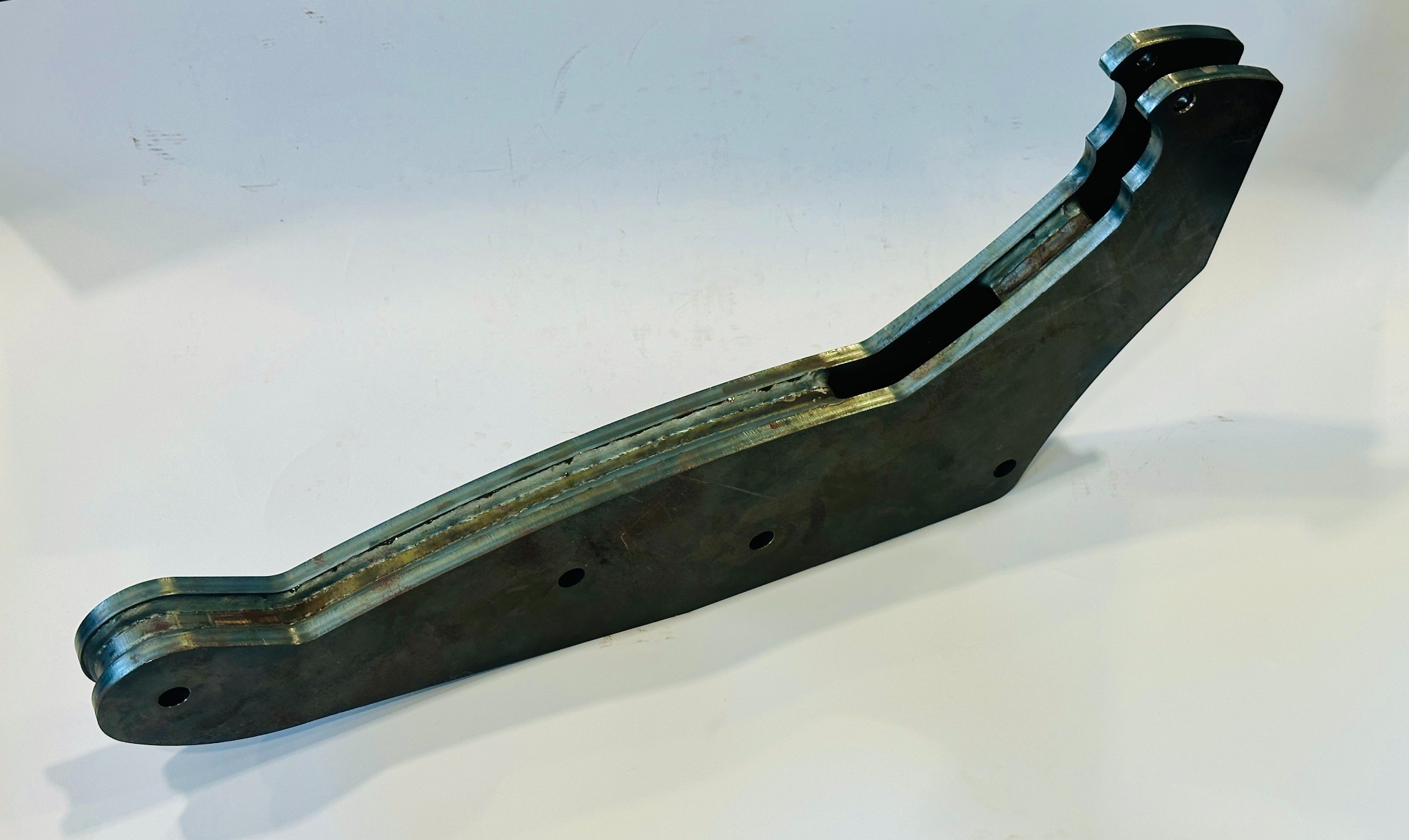 Metalowy element konstrukcyjny z widocznymi spawami i otworami, leżący na białym tle. Detal wykonany z kilku warstw blachy, z wyraźnymi śladami obróbki.