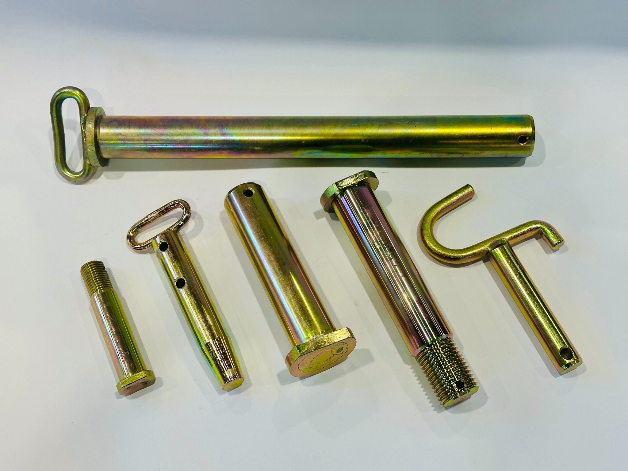Zestaw sześciu metalowych sworzni w odcieniach złota i srebra, o różnych kształtach i rozmiarach, leżących na białym tle. Widoczne gwinty i otwory.
