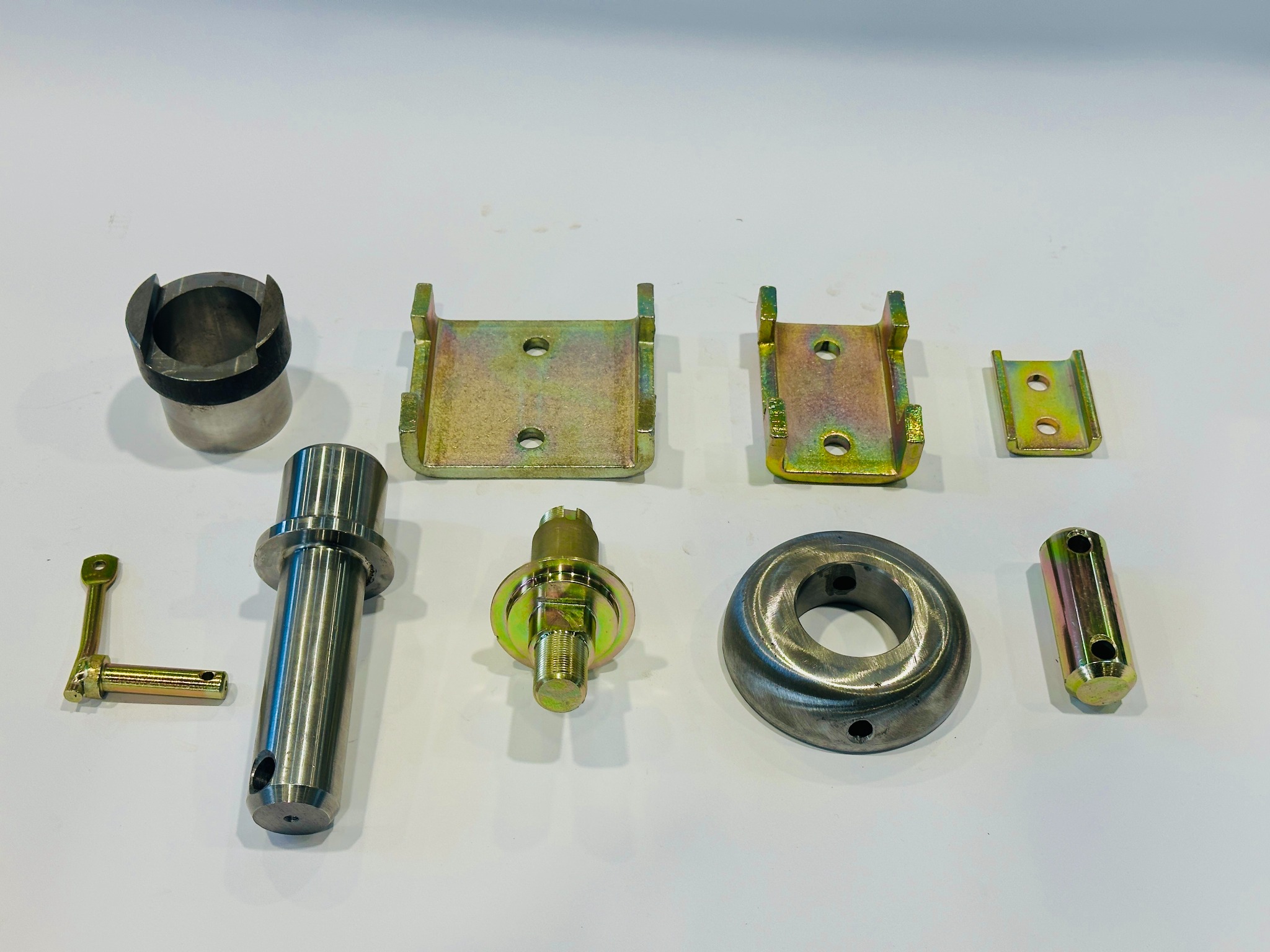 Precyzyjnie wykonane, metalowe elementy z żółtym ocynkiem, ułożone na białym tle. Widoczne różne kształty i rozmiary, w tym tuleje, sworznie i mocowania.